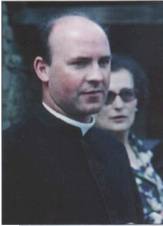 Priest standing with woman in background.